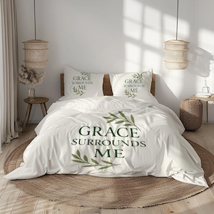 May include: White bedding set with the text "GRACE SURROUNDS ME" in dark green, and olive branch designs. The set includes a duvet cover and two pillowcases. The bed is on a round, woven rug.