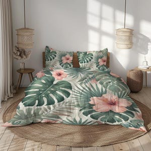 Puede incluir: Un juego de cama de temática tropical con fondo blanco y un estampado a gran escala de hojas verdes de monstera y flores de hibisco rosas.