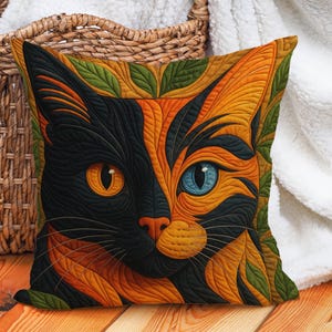 Puede incluir: Cojín decorativo cuadrado con la cara de un gato estilizada. La cara del gato está dividida, un lado negro y el otro naranja, con un ojo azul y otro naranja. El cojín está bordeado por hojas verdes y descansa sobre una superficie de madera.