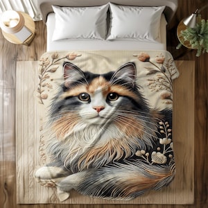 Op de afbeelding: Een beige sprei met een gedetailleerde illustratie van een lapjeskat. De kat heeft bruin, zwart en wit haar en is omgeven door bloemmotieven. De deken ligt op een bed met witte kussens.