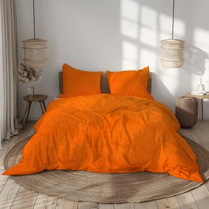 May include: Bright orange bed set featuring a duvet cover and two matching pillowcases. The bedding is arranged on a wooden bed frame, with a neutral-toned rug beneath. The room is decorated with hanging woven lamps and a small side table.