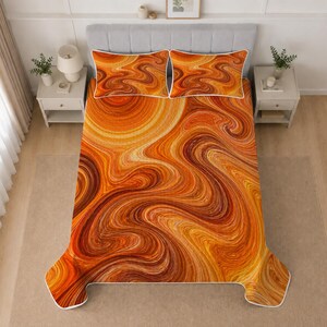 May include: A bed set featuring a quilt and two matching pillow shams with a vibrant orange, yellow, and brown marbled swirl pattern. The bedding has a white piped edge detail and is displayed on a bed with a light gray upholstered headboard.