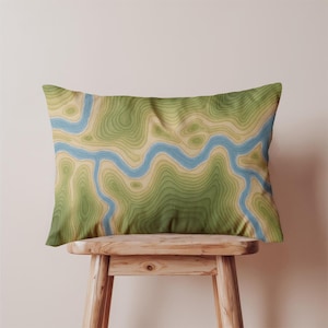 May include: A rectangular pillow with a topographic map design. The pillow features a blue river winding through green and tan contour lines, creating a landscape pattern. The pillow is resting on a wooden stool.