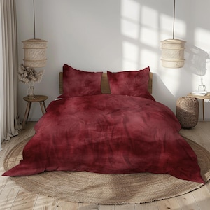 Può includere: Un letto con un copripiumino bordeaux e due federe abbinate. La biancheria da letto ha un aspetto morbido e strutturato. Il letto è appoggiato su un tappeto rotondo intrecciato. Due lampade a sospensione intrecciate sono appese sopra il letto.