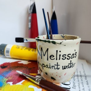 May include: A white ceramic paint water cup with black lettering that says "Melissa's paint water". The cup has a speckled design and is filled with paint brushes.
