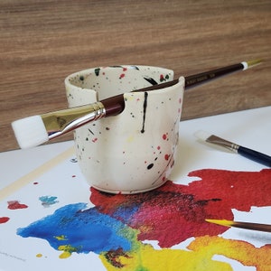 May include: White ceramic paintbrush holder with black, red, and green splatters. A paintbrush is resting in the holder.
