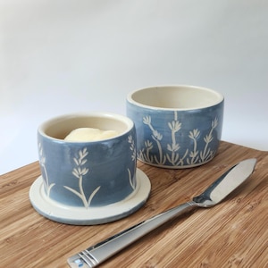 May include: Two blue ceramic butter dishes with white floral designs. One dish contains butter. A stainless steel butter knife rests on a wooden cutting board. The dishes have a cylindrical shape with a lid.