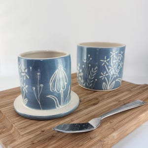 May include: A blue ceramic butter crock set consisting of a base with a lid and a separate matching container, both featuring white hand-carved floral patterns. A silver-toned metal butter knife is placed in the foreground.