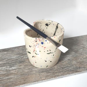 May include: A white ceramic paintbrush holder with a speckled design in shades of blue, green, red, and yellow. A blue and white paintbrush is resting in the holder.