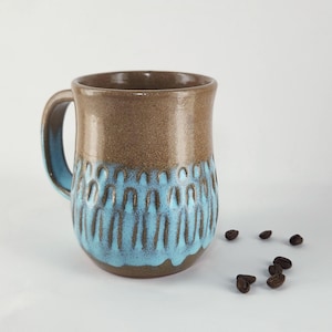 Puede incluir: Una taza de cerámica con una sección superior marrón lisa y una sección inferior texturizada con un esmalte azul brillante. La taza tiene un asa curva con un acabado interior azul. Varios granos de café están esparcidos cerca de la base.