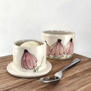 May include: A ceramic butter crock with a lid and a matching bowl, both featuring hand-painted pink coneflower designs with green stems. A small silver-toned metal spoon rests beside the pottery on a wooden surface, completing the set.