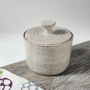 May include: A cylindrical ceramic jar with a matching lid, featuring a light cream-colored glaze with dark brown speckles. The exterior of the jar and the top of the lid are decorated with fine, horizontal concentric grooves and a knob handle.