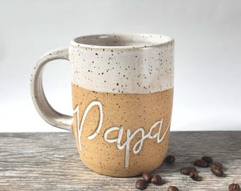 May include: A ceramic mug with a speckled white upper section and a tan lower section. The word Papa is embossed in white cursive script on the front. The mug features a curved handle and is accompanied by several scattered dark coffee beans.