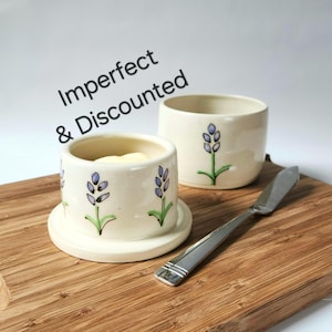 May include: Cream-colored ceramic butter dish with a matching lid, decorated with lavender sprigs. A stainless steel butter knife rests on a wooden cutting board. The text "Imperfect & Discounted" is in the upper left corner.