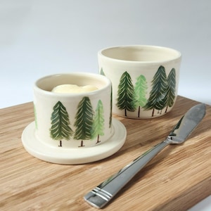 May include: A cream-colored butter dish and lid with a forest design in shades of green. The butter dish sits on a wooden cutting board next to a stainless steel butter knife. The butter dish contains butter.