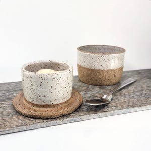 May include: Two ceramic ramekins with speckled white glaze and a natural cork base. One ramekin sits on a matching coaster, and the other is next to a spoon. The ramekins are on a wooden surface.