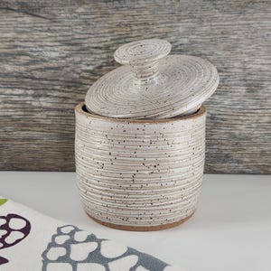 May include: A cylindrical ceramic canister with a speckled off-white glaze and horizontal ridged texture. The matching lid features a central knob handle and rests slightly askew on top of the container, revealing the interior rim.