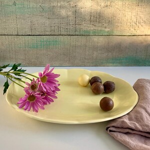 May include: A pale yellow, scalloped-edge ceramic serving platter. The platter holds several chocolate truffles and a single white chocolate. A bouquet of pink flowers with yellow centers rests on the platter's edge. A pink cloth is on the right.
