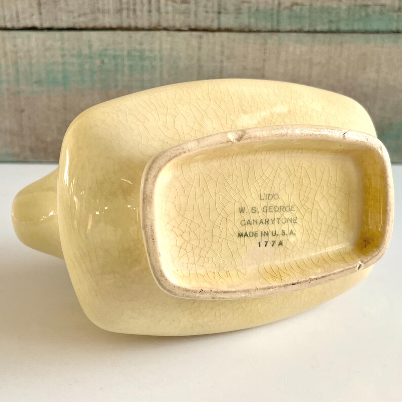 May include: A vintage, pale yellow ceramic pitcher with a spout and handle. The bottom of the pitcher features the text "Lido W.S. George Canarytone Made in U.S.A. 177A". The pitcher has a crackle glaze finish.