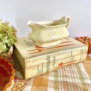 May include: A pale yellow ceramic gravy boat with a handle and spout, resting on a vintage cookbook. The cookbook is titled "Encyclopedic Cookbook" and features illustrations of utensils. The scene is set on a checkered tablecloth.