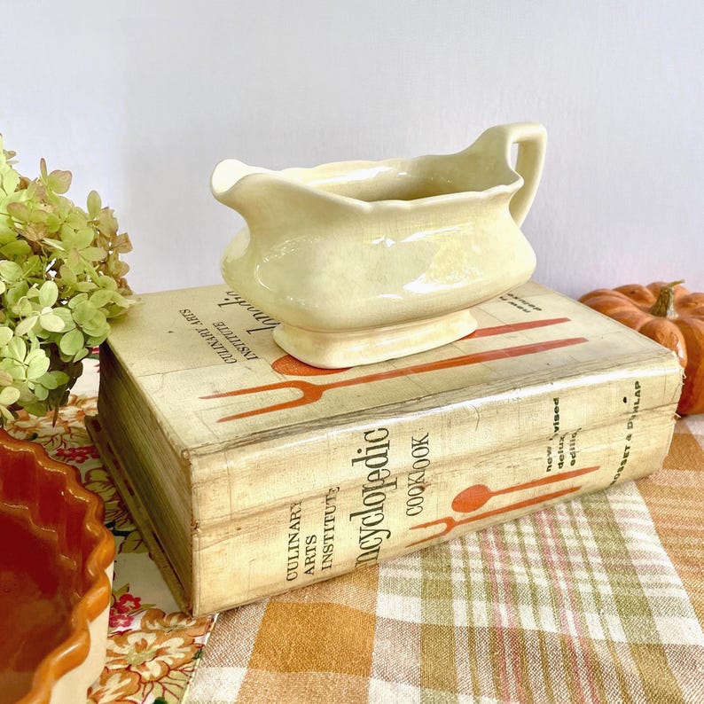 May include: A pale yellow ceramic gravy boat with a handle and spout, resting on a vintage cookbook. The cookbook is titled "Encyclopedic Cookbook" and features illustrations of utensils. The scene is set on a checkered tablecloth.