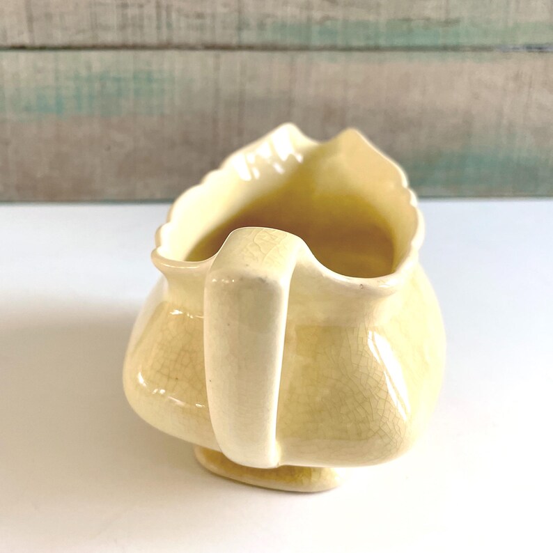 May include: A vintage, pale yellow ceramic gravy boat with a scalloped rim and a curved handle. The gravy boat has a small footed base and a crackle glaze finish, giving it a classic, aged appearance.