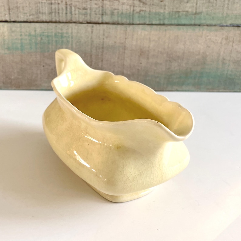 May include: A vintage, cream-colored ceramic gravy boat with a scalloped rim and a handle. The gravy boat has a rounded body and a slightly flared spout. The interior is a darker shade of cream. The piece is likely from the mid-20th century.