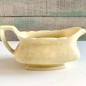 May include: A pale yellow ceramic gravy boat with a handle. The boat has a wide, curved rim and sits on a small, rectangular base. The surface has a crackle glaze effect.