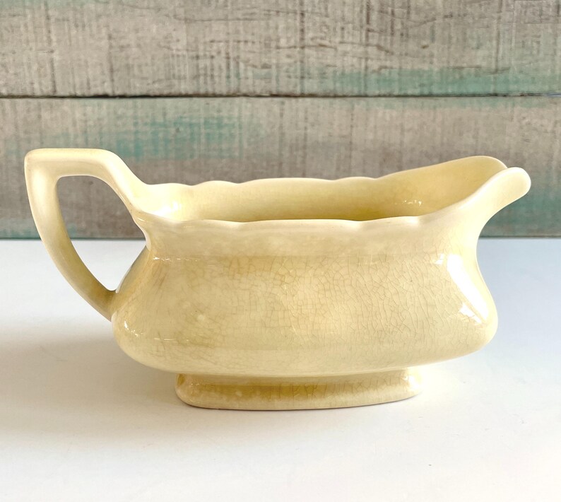 May include: A pale yellow ceramic gravy boat with a handle. The boat has a wide, curved rim and sits on a small, rectangular base. The surface has a crackle glaze effect.