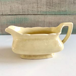 May include: A pale yellow ceramic gravy boat with a handle. The boat has a curved spout and sits on a small, rectangular base. The handle is arched and attached to the side of the boat. The gravy boat is a vintage style.