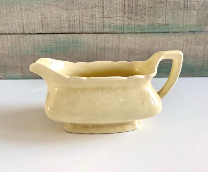 May include: A pale yellow ceramic gravy boat with a handle. The boat has a curved spout and sits on a small, rectangular base. The handle is arched and attached to the side of the boat. The gravy boat is a vintage style.