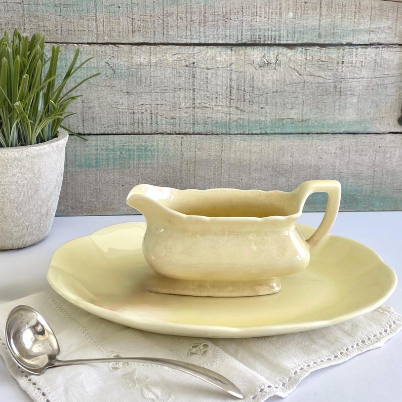 May include: A vintage pale yellow gravy boat with a matching plate. The gravy boat has a handle and sits on a raised base. A silver ladle rests on a white embroidered napkin. The set is on a white surface.