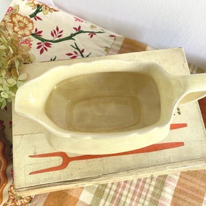 May include: A pale yellow ceramic gravy boat with a scalloped rim and a handle. The gravy boat is sitting on a vintage book with a red fork illustration. A floral patterned fabric is in the background.