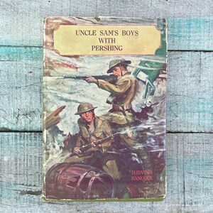 May include: A vintage hardcover book titled Uncle Sam's Boys with Pershing by H. Irving Hancock. The cover features a color illustration of two soldiers in tan uniforms and helmets holding rifles in a battlefield scene with a wooden barrel in the foreground.