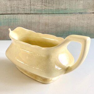 May include: A pale yellow ceramic gravy boat with a handle. The boat has a rectangular base and a scalloped rim. The handle is a simple loop shape. The surface has a crackle glaze.