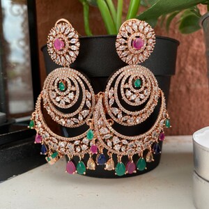 May include: A pair of gold tone earrings with a large, round, multi-layered design. The earrings are adorned with clear and green stones, as well as pink and blue accents.