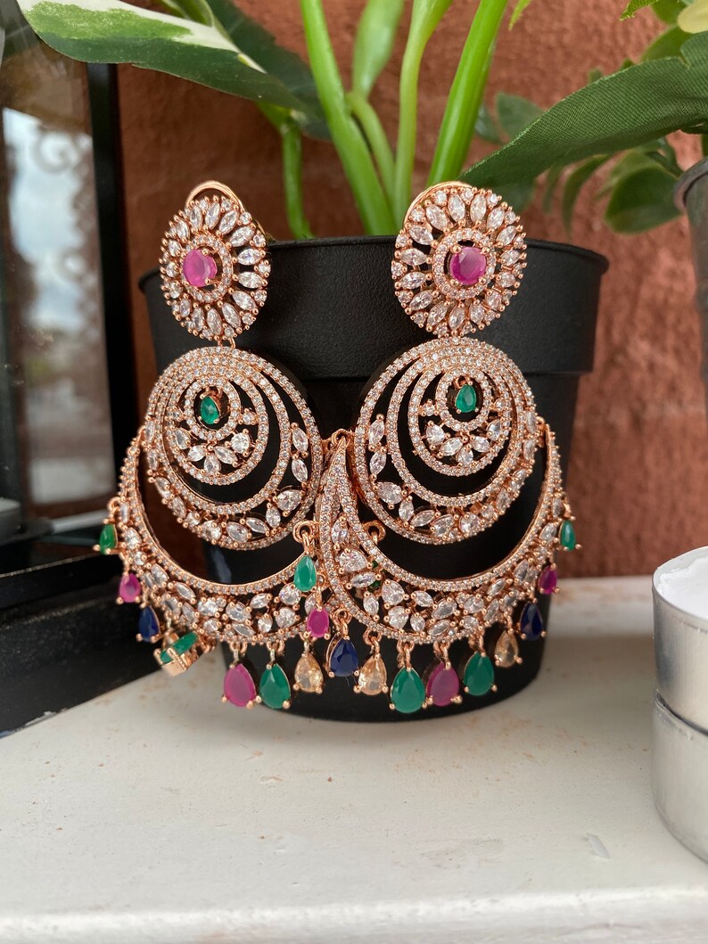 May include: A pair of gold tone earrings with a large, round, multi-layered design. The earrings are adorned with clear and green stones, as well as pink and blue accents.