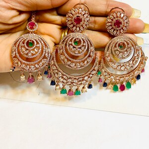 May include: A pair of gold-tone earrings with a multi-layered design. The earrings feature a large, round, openwork design with smaller circles and teardrop-shaped stones. The earrings are adorned with clear, white, and colored stones, including red, green, and blue.