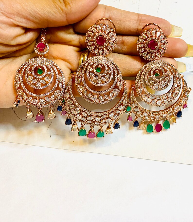 May include: A pair of gold-tone earrings with a multi-layered design. The earrings feature a large, round, openwork design with smaller circles and teardrop-shaped stones. The earrings are adorned with clear, white, and colored stones, including red, green, and blue.