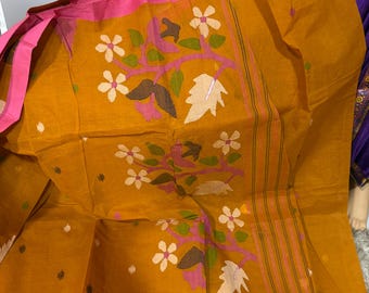 May include: A mustard yellow fabric featuring a pink border and a repeating pattern of pink, white, and green floral motifs with brown leaves. The textile displays a woven texture with thin vertical stripes along one side and small scattered dots.