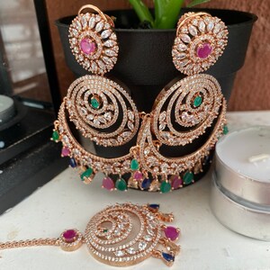 May include: A set of gold-tone earrings and a matching nose ring. The earrings feature a large, circular design with smaller circles and pink, green, and blue stones. The nose ring has a similar design with a smaller circle and pink stones.