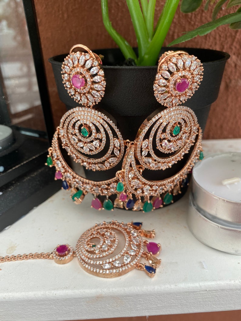 May include: A set of gold-tone earrings and a matching nose ring. The earrings feature a large, circular design with smaller circles and pink, green, and blue stones. The nose ring has a similar design with a smaller circle and pink stones.