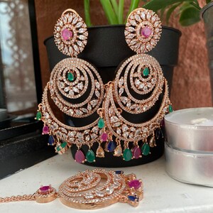 May include: A set of gold-tone earrings and a matching nose ring with a circular design. The earrings have multiple layers of sparkling stones and dangle with pink, green, and blue gems. The nose ring has a similar design with a smaller size and a single pink gem.