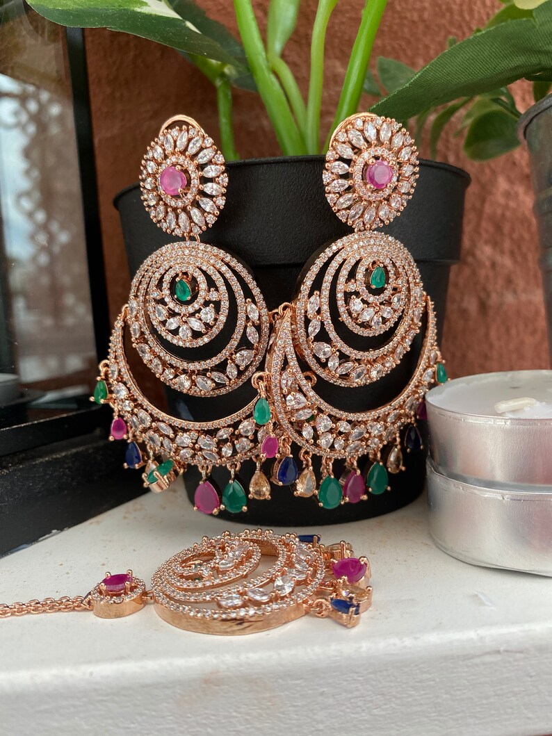 May include: A set of gold-tone earrings and a matching nose ring with a circular design. The earrings have multiple layers of sparkling stones and dangle with pink, green, and blue gems. The nose ring has a similar design with a smaller size and a single pink gem.