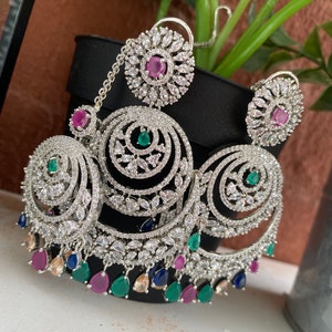 May include: Silver earrings with a circular design, featuring clear stones and pink, green, and blue accents. The earrings have a dangling chain with a circular pendant.