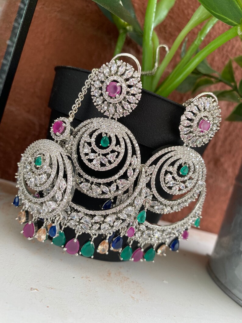 May include: Silver earrings with a circular design, featuring clear stones and pink, green, and blue accents. The earrings have a dangling chain with a circular pendant.