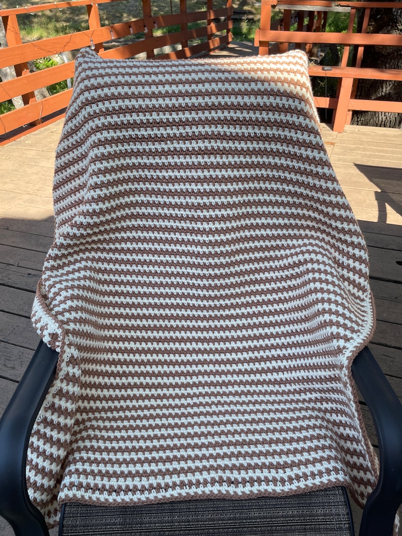 May include: A brown and white striped crocheted throw blanket is draped over a black chair on a wooden deck.