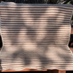 May include: A brown and white striped crocheted throw blanket with a textured pattern. The blanket is folded and resting on a wooden bench.