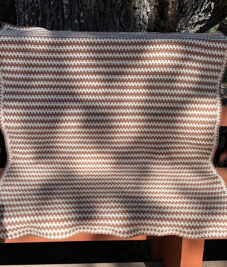 May include: A brown and white striped crocheted throw blanket with a textured pattern. The blanket is folded and resting on a wooden bench.
