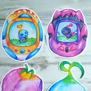 May include: Four colorful watercolor stickers. Two depict retro handheld game consoles with cartoon characters inside. The other two are a pink strawberry and a blue and green onion-shaped design.